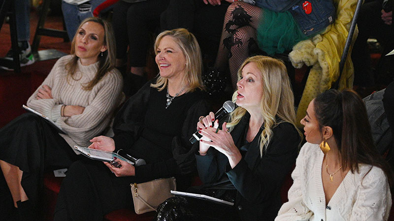 Four women sitting in a row judging a contest, one holding a microphone and speaking