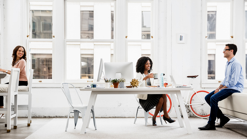 marquee-office-people-800x450 Man with laptop on table and woman with coffee cup seated at table discuss topic while woman at another table turns and smiles.