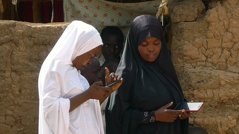 Two women holding phones are looking down at their hands in a marketplace.