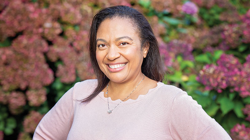 Woman entrepreneur wearing pink sweater standing in front of flowers and looking at the camera.