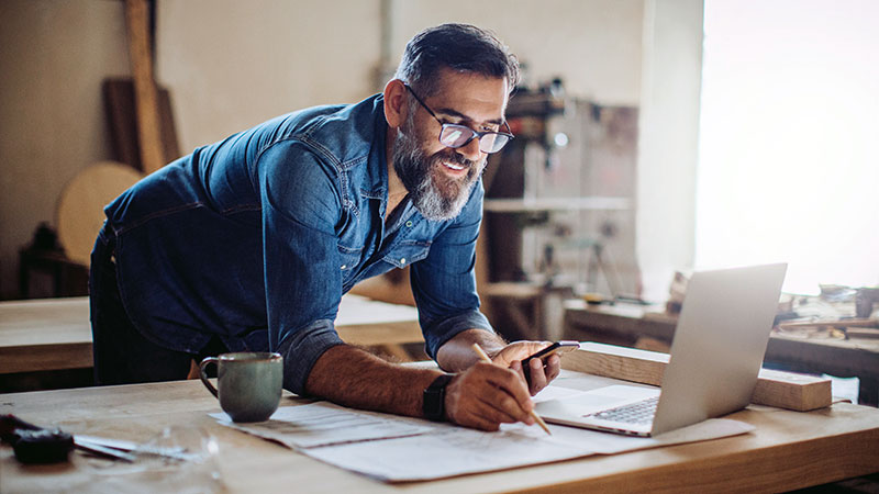 Man leaning over a desk looking a business papers, a phone and a laptop, running a small business.