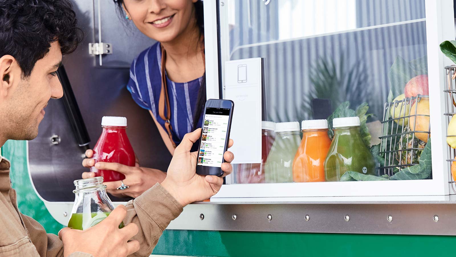 Man looking at his mobile phone while standing near a food truck and woman smiling and serving him some sauce.
