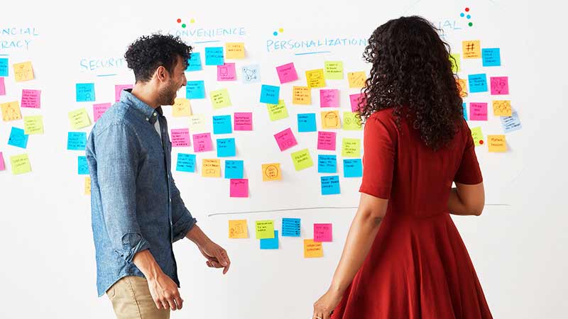 A pair of Visa employees review a white board full of sticky notes. A pair of Visa employees review a white board full of sticky notes.