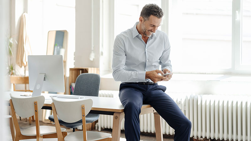 Man looking at his smartphone while sitting at the edge of his desk.