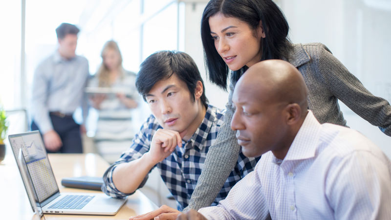 Three people in an office setting looking at a spread sheet on a laptop
