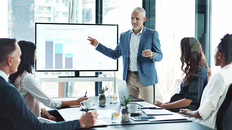 A business presentation where a man points to a display featuring graphs.