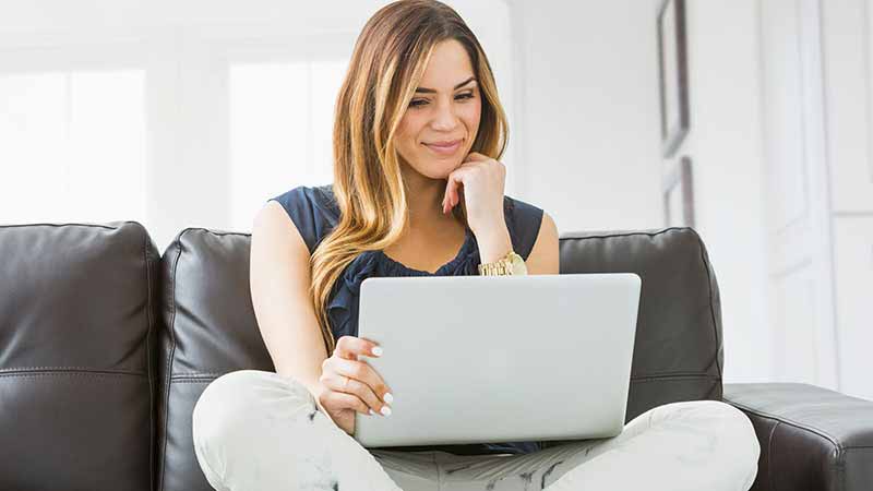 A smiling woman on a couch looks at a laptop.