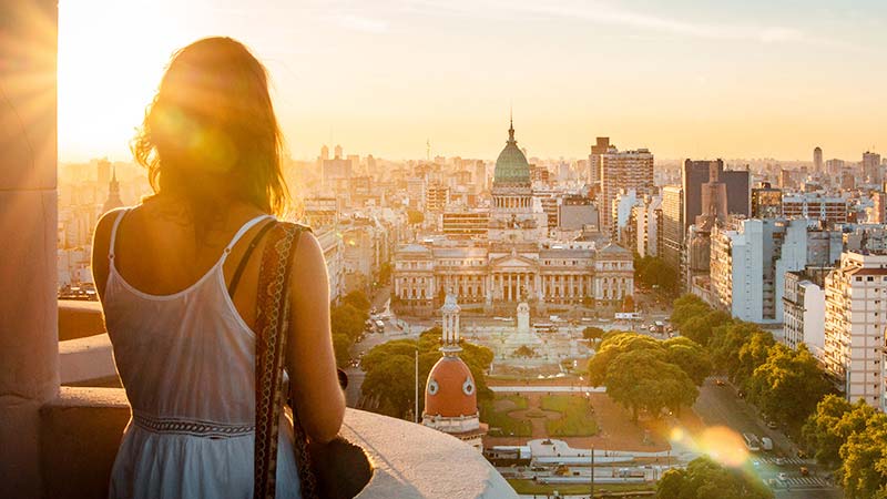 A traveler on a balcony looks out over the city of Buenos Aires at sunset.