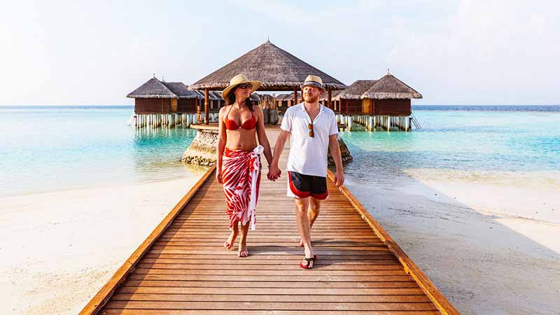 A couple hold hands as they stroll away from a cluster of overwater huts.