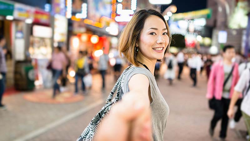 A smiling young Asian woman leads her companion by the hand through a busy shopping district.