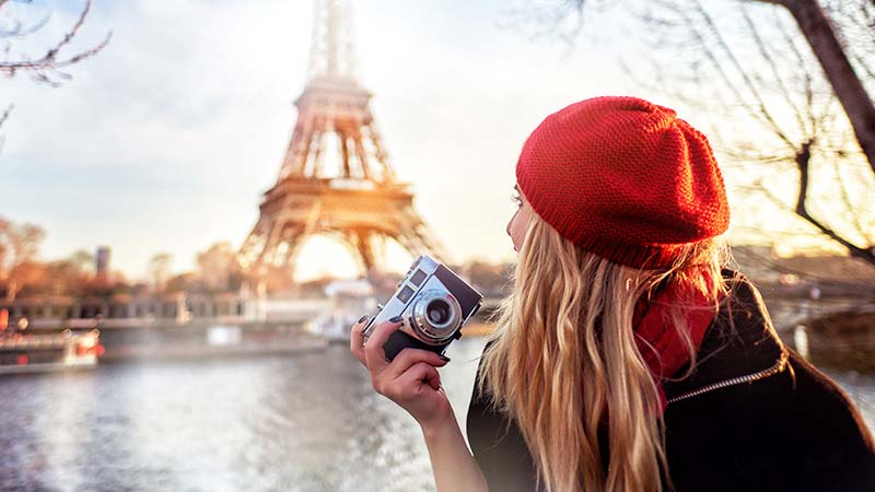 A young woman holding a camera in front of the Eiffel Tower in Paris.